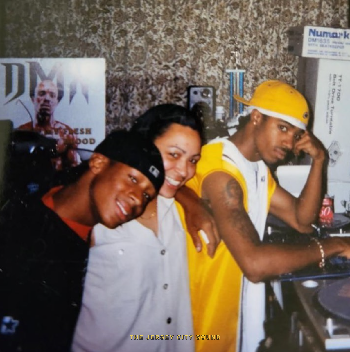 Three friends at a home turntable setup with a DMX poster and Numark boxes