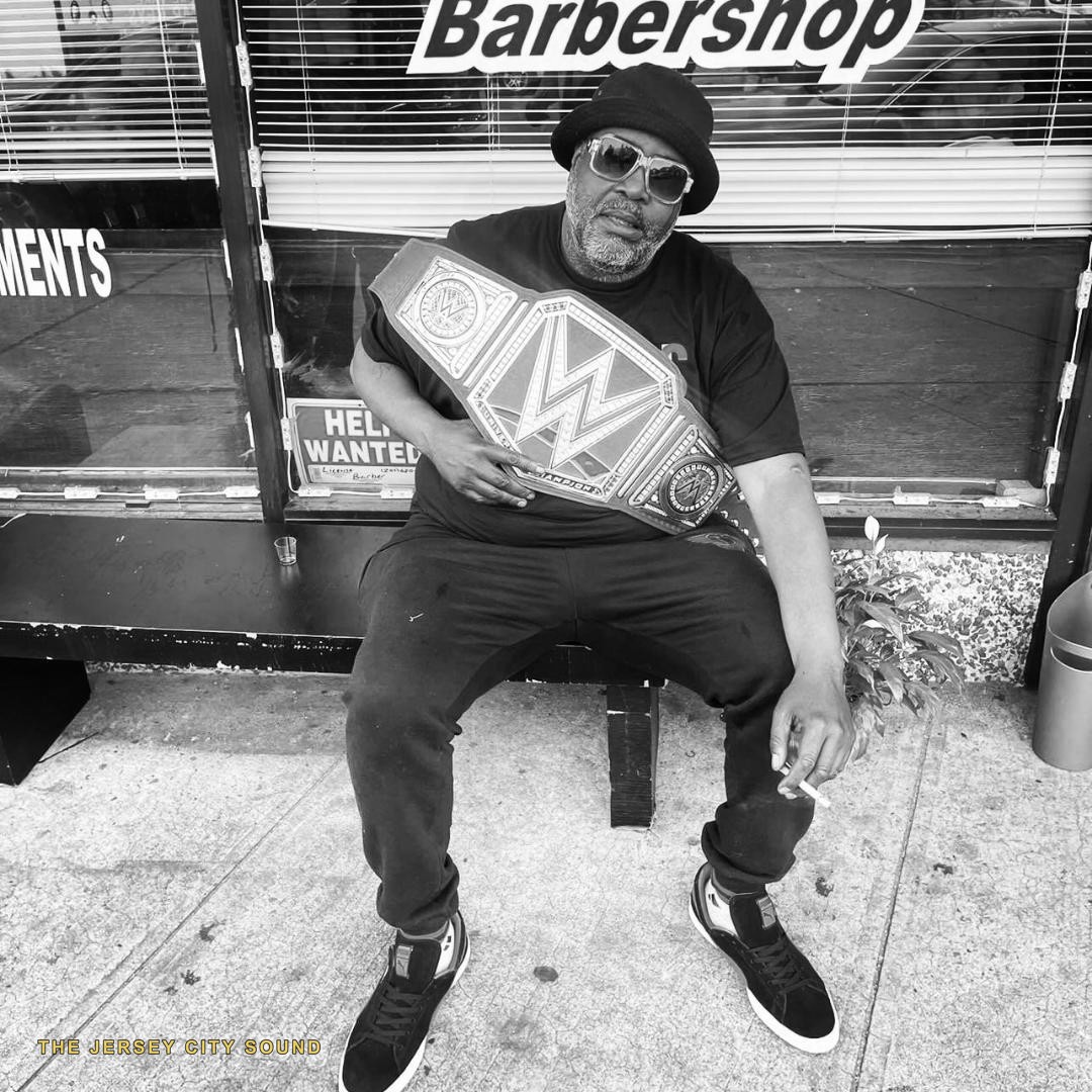 DJ Flash seated outside a barbershop holding a championship belt