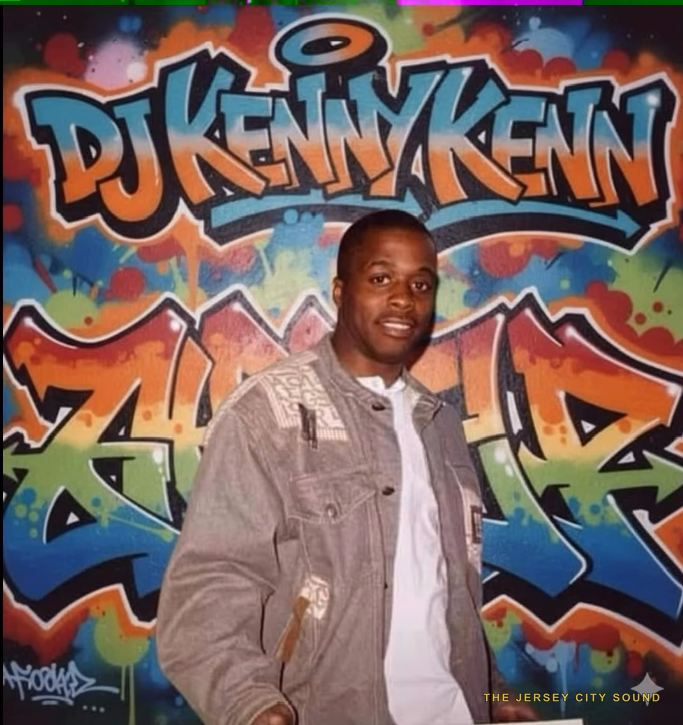 DJ Kenny Kenn smiling in front of a graffiti mural bearing his name
