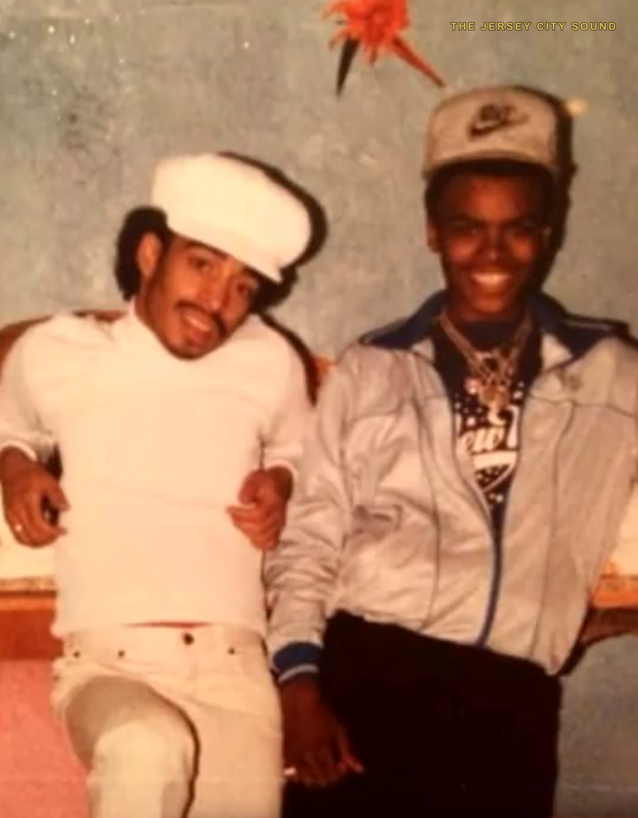 Two men seated together at a gathering, one in a white sailor cap, one in a Nike cap