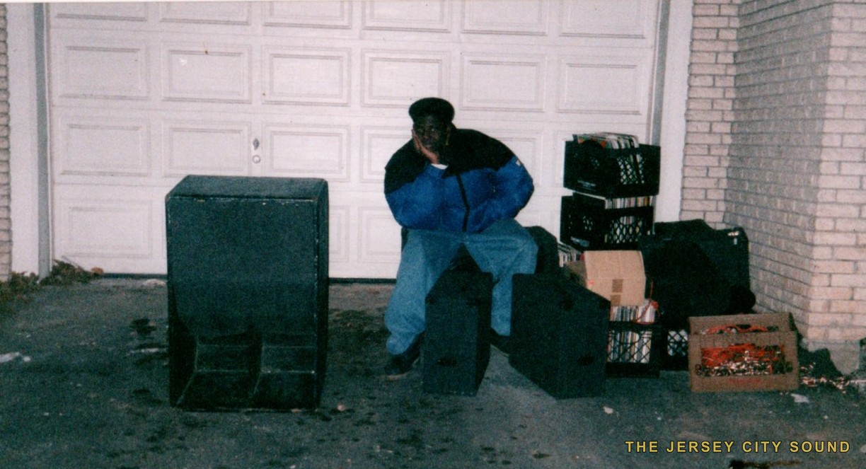 A young Mista Quietman seated among speaker cabinets and milk crates of records